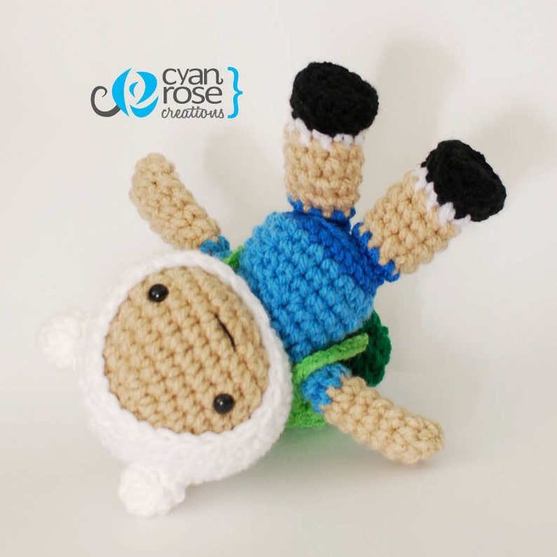 May include: Crocheted Finn the Human from Adventure Time, wearing a blue and green shirt and white hat. He is lying on his back with his legs in the air.