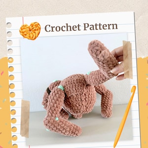 Può includere: Un modello di coniglio all'uncinetto marrone con la scritta "Crochet Pattern". Il coniglio ha accenti verde chiaro sugli arti. Una mano tiene uno degli arti del coniglio. Un uncinetto è nell'immagine.