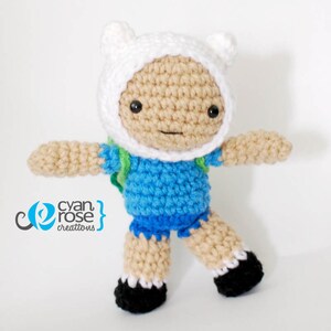 May include: A crocheted plush toy of Finn the Human from the animated series Adventure Time. Finn is wearing a blue shirt, green backpack, and white hat with ears.