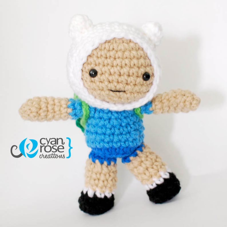 May include: A crocheted plush toy of Finn the Human from the animated series Adventure Time. Finn is wearing a blue shirt, green backpack, and white hat with ears.