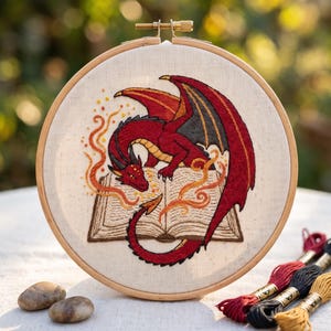May include: A circular wooden embroidery hoop containing a fabric panel with a hand-stitched design of a red dragon with black wings perched over an open book. The dragon is surrounded by swirling orange and yellow flame-like embroidery thread accents.