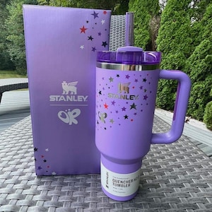 May include: A purple Stanley Quencher H2.0 Flowstate tumbler with a handle and a matching box. The tumbler and box feature the Stanley logo and a pattern of stars in various colors. The tumbler has a straw and the text "Quencher H2.0 Tumbler".