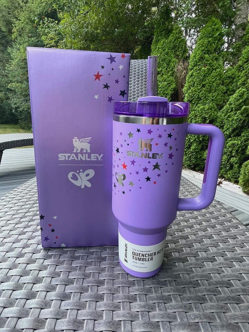 May include: A purple Stanley Quencher H2.0 Flowstate tumbler with a handle and a matching box. The tumbler and box feature the Stanley logo and a pattern of stars in various colors. The tumbler has a straw and the text "Quencher H2.0 Tumbler".