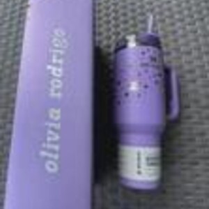 May include: A lavender insulated tumbler with a handle and straw, next to a matching box. The tumbler features a pattern of small stars. The box has the text "olivia rodrigo" printed on it.