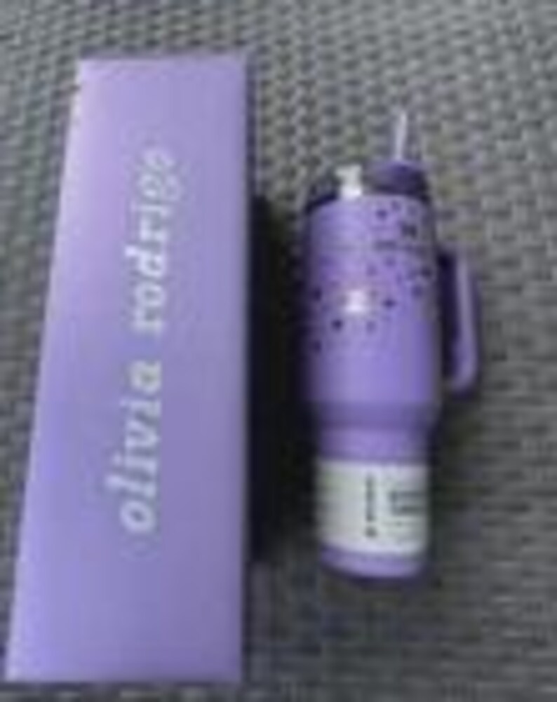 May include: A lavender insulated tumbler with a handle and straw, next to a matching box. The tumbler features a pattern of small stars. The box has the text "olivia rodrigo" printed on it.