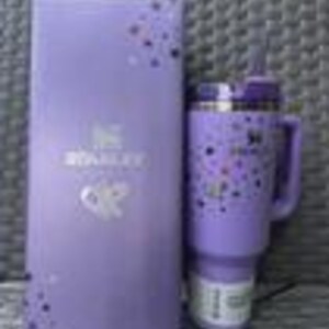 May include: A purple Stanley Quencher H2.0 FlowState tumbler with a handle and straw, next to a matching box. The tumbler features a speckled design. The box has the Stanley logo and text. The tumbler is approximately 9 inches tall.