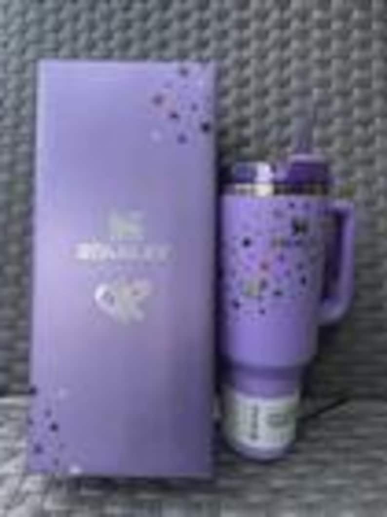 May include: A purple Stanley Quencher H2.0 FlowState tumbler with a handle and straw, next to a matching box. The tumbler features a speckled design. The box has the Stanley logo and text. The tumbler is approximately 9 inches tall.