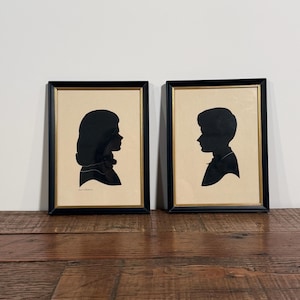 May include: Two framed silhouette portraits featuring black profiles of individuals on a cream-colored background. Each portrait is encased in a rectangular black frame with a thin gold inner border, displaying a side view of a head and shoulders.
