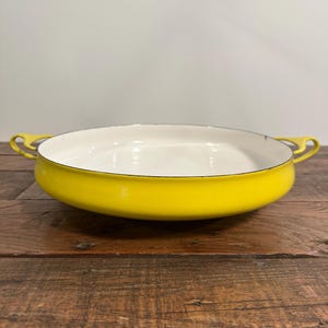 May include: A round, shallow baking dish with a vibrant yellow exterior and a smooth, white interior. The vessel features two integrated yellow handles on opposite sides and a thin, dark rim around the top edge of the circular cooking surface.