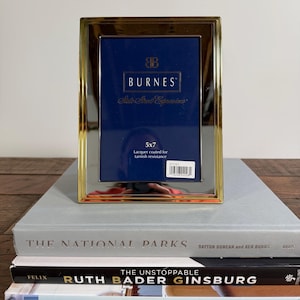 May include: A rectangular gold-toned metal picture frame with a polished finish. The frame contains a blue display insert with the text BURNES, State Street Expressions, 5x7, and Lacquer coated for tarnish resistance, along with a barcode sticker.
