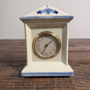 May include: A cream-colored ceramic mantel clock with a blue floral motif on the roof-shaped top. The clock face is white with black numerals, a gold-tone circular bezel, and the brand name Zuriol printed in the center. The ceramic has a crackle finish.