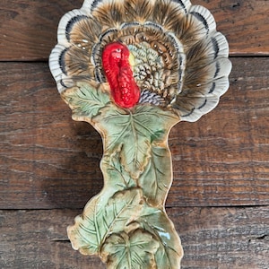 May include: A ceramic spoon rest shaped like a turkey with a fanned tail featuring brown, white, and black feathers. The turkey has a bright red head and neck, resting on a base of sculpted green leaves with a glossy finish and visible texture.
