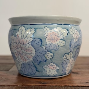 May include: A round ceramic planter featuring a light blue background with a repeating pattern of white and soft pink chrysanthemum flowers and blue leaves. The bowl has a wide, smooth white rim and a tapered base with a glossy finish.