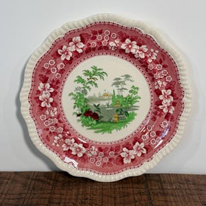May include: A round ceramic plate with a scalloped edge featuring a cream-colored center with a green landscape illustration of trees and a building. The border is decorated with a deep pink floral pattern on a lighter pink background.