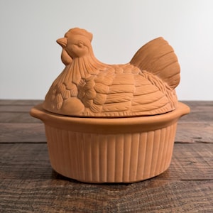 May include: A terracotta lidded casserole dish featuring a sculpted chicken on the lid. The base is round with vertical ribbed sides and two small handles. The lid is shaped like a sitting hen with detailed feather textures and a comb.
