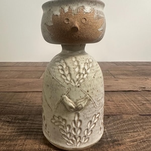 May include: A ceramic vase featuring a stylized figure with a round head, a pointed nose, and two circular eyes. The body is off-white with brown speckles and carved wheat-like patterns, showing two hands clasped together at the center.