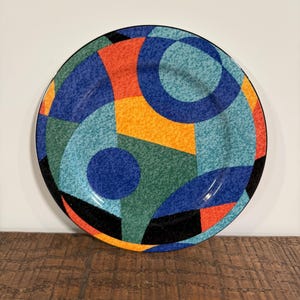 May include: A round ceramic plate featuring a vibrant geometric pattern with overlapping circles, triangles, and segments in shades of blue, teal, yellow, orange, and black. The surface has a textured, speckled finish throughout the colorful design.
