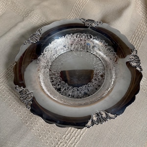 May include: A round silver-toned metal bowl with a scalloped edge and a polished finish. The interior features an intricate embossed floral and scrollwork pattern around the centre, while the rim is accented with four decorative metal filigree motifs.