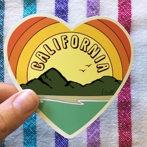 Op de afbeelding: Een hartvormige sticker met een retro zonsondergangontwerp. De sticker heeft het woord "CALIFORNIA" in een geel, oranje en rood kleurenschema. De sticker bevat ook een afbeelding van bergen en een strand.