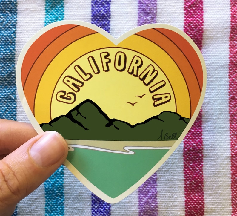Op de afbeelding: Een hartvormige sticker met een retro zonsondergangontwerp. De sticker heeft het woord "CALIFORNIA" in een geel, oranje en rood kleurenschema. De sticker bevat ook een afbeelding van bergen en een strand.