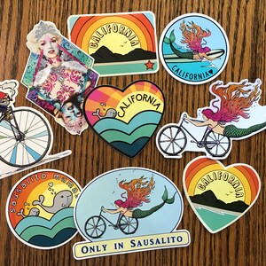 May include: A set of nine colorful stickers featuring various designs, including a mermaid riding a bicycle, a mermaid on a surfboard, a whale, a gnome riding a penny-farthing bicycle, and a heart with the words "California" inside. The stickers are all on a wooden surface.