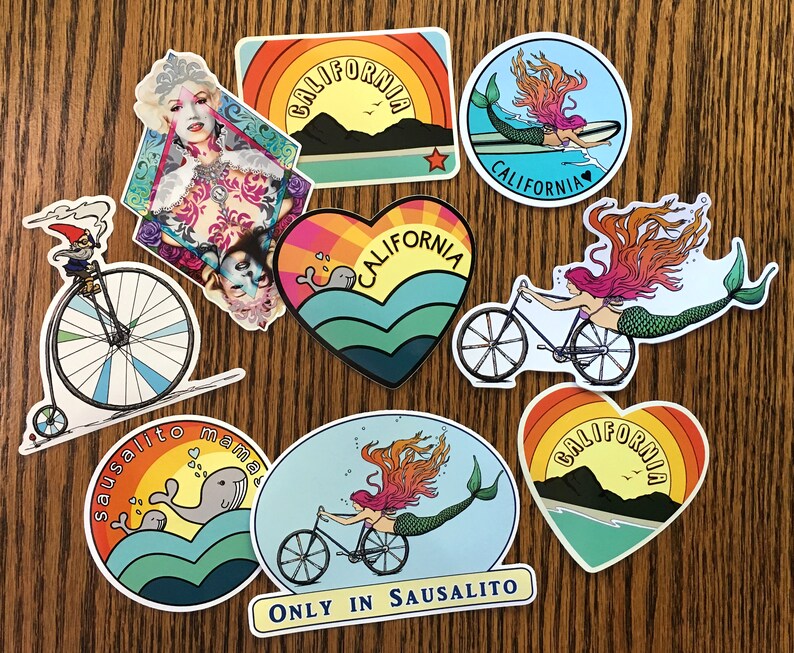 May include: A set of nine colorful stickers featuring various designs, including a mermaid riding a bicycle, a mermaid on a surfboard, a whale, a gnome riding a penny-farthing bicycle, and a heart with the words "California" inside. The stickers are all on a wooden surface.