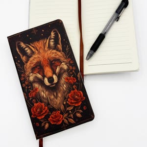 May include: A journal with a fox illustration on the cover, surrounded by red roses and foliage. The fox has orange and brown fur, with a black pen and a lined notebook in the background.
