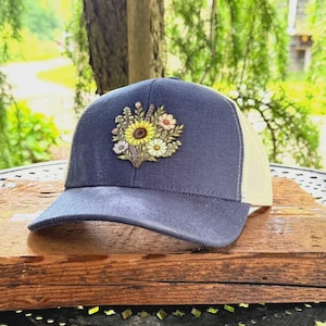May include: A navy blue baseball cap with a cream-colored mesh back panel. The front of the cap features a detailed, multi-colored embroidered patch of a floral bouquet, including a large yellow sunflower, white daisies, and small purple flowers.