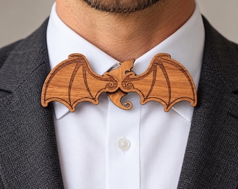 May include: A wooden bow tie shaped like a pair of bat wings with a central bat body silhouette. The accessory features a natural light brown wood grain finish with dark engraved lines detailing the wing structure and is worn on a white shirt.