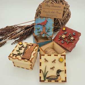 May include: Four square wooden boxes with interlocking edges, each featuring layered wood cutouts of birds and nature scenes. Designs include a red cardinal, a woodpecker, small birds on a branch, and a loon with dragonflies. Each box lid displays a sun.