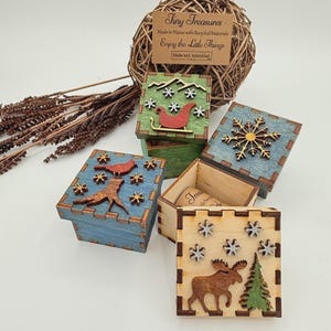 May include: Four square wooden boxes with laser-cut designs. One green box features a red sleigh and mountains. Two blue boxes show a snowflake and a cardinal on a stump. One natural wood box displays a moose and tree. All have snowflake accents.
