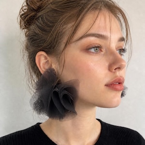 May include: Black tulle earrings in a floral design. The earrings are large and eye-catching, adding a dramatic flair. The earrings are worn with a black top.