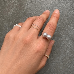 May include: A hand wearing several rings. The rings include a delicate pearl band, two thin gold bands, a ring with two large pearls, and a small band with small stones. The rings are displayed against a gray background.