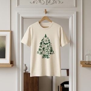 Może przedstawiać: Kremowy t-shirt z krótkim rękawem o nakrapianej fakturze. Na środku znajduje się duża, ciemnozielona grafika w kształcie sosny, złożona z różnych ikon outdoorowych, w tym sasquatcha, łosia, drzew, namiotu i ogniska.