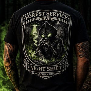 May include: Black t-shirt with a graphic featuring a forest scene, an alien, and the text "FOREST SERVICE NIGHT SHIFT APPALACHIAN DIVISION". The alien has glowing green eyes and is smoking.