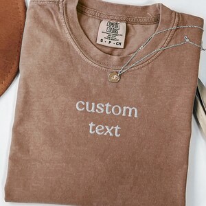 May include: A brown short-sleeved t-shirt with a crew neckline and the words custom text embroidered in white thread on the chest. The shirt features a Comfort Colors brand label at the inner neck and is paired with a thin silver-toned necklace.