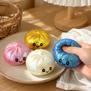 Può includere: Quattro giocattoli rotondi e morbidi a forma di gnocco con finiture glitterate in rosa, giallo, bianco e blu. Ogni giocattolo presenta un semplice viso stampato con due occhi neri e una piccola bocca sorridente. Il giocattolo blu viene schiacciato.