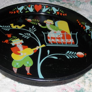 May include: A black metal tray with a colorful folk art design. The design features a couple, a tree, and birds. The tray is decorated with red, yellow, blue, green, and white paint.