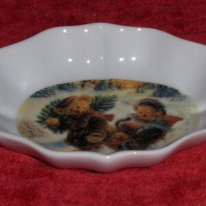 May include: A small, white, scalloped-edge dish with a winter scene featuring two teddy bears in a snowy landscape. One bear carries a pine branch, and the other is sledding. The dish is likely ceramic.