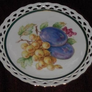 May include: A white porcelain plate with a green and gold rim and a lace-like edge. The plate features a painted design of plums and grapes.