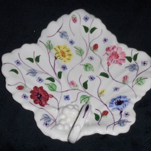 May include: White ceramic leaf-shaped dish with a floral design in shades of pink, red, yellow, blue, and green. The dish has a raised handle in the shape of a bunch of grapes.