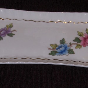 May include: A white rectangular ceramic dish with a gold trim and a floral design. The dish has a scalloped edge and features pink, blue, and purple flowers.