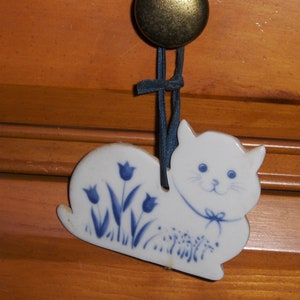 May include: A white ceramic cat figurine with blue floral details hanging from a blue ribbon. The cat is sitting with its head tilted and has a sweet expression.
