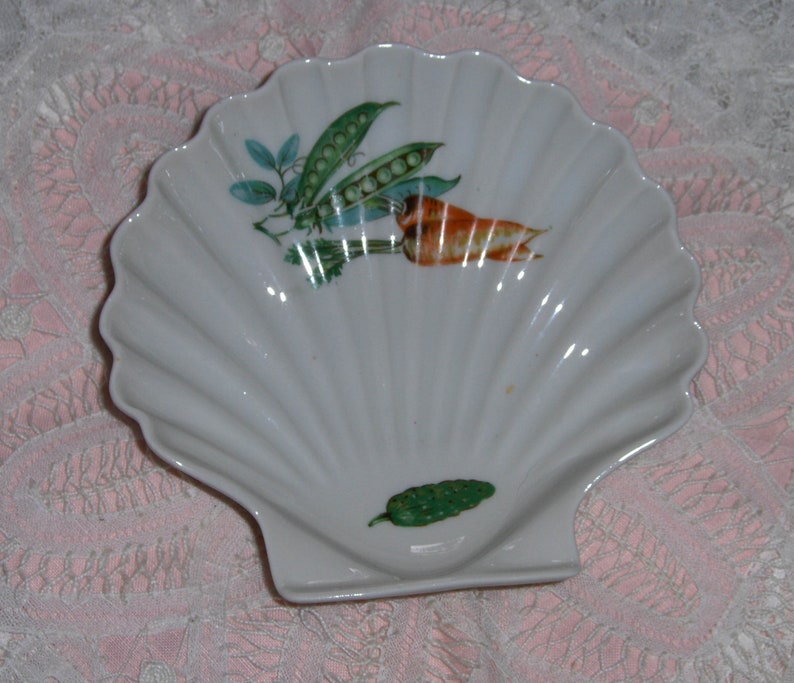K&ouml;nnte beinhalten: Wei&szlig;e Porzellanschale in Muschelform mit einem Design aus gr&uuml;ner Erbsenkapsel, orangefarbener Karotte und gr&uuml;nem Blatt.
