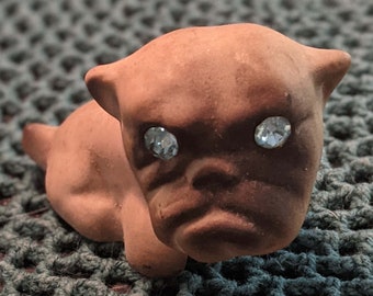 Peut inclure: Petite figurine en céramique marron représentant un chien, avec des yeux en strass bleu clair. Le chien est couché, la tête relevée, le pelage plus foncé autour du museau. La figurine est posée sur une surface crochetée verte.