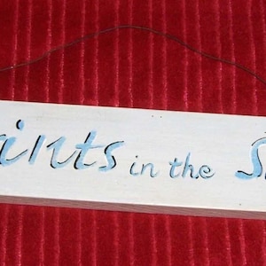 May include: A rectangular wooden sign with rounded edges, painted white with the words "footprints in the sand" in blue script. A thin wire is attached for hanging. The sign is against a red background.