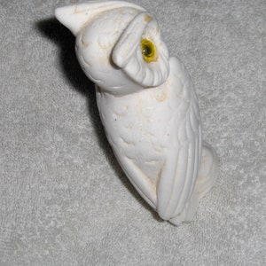 Puede incluir: Figurita de b&uacute;ho de cer&aacute;mica blanca con un ojo amarillo. El b&uacute;ho est&aacute; posado sobre una superficie blanca.