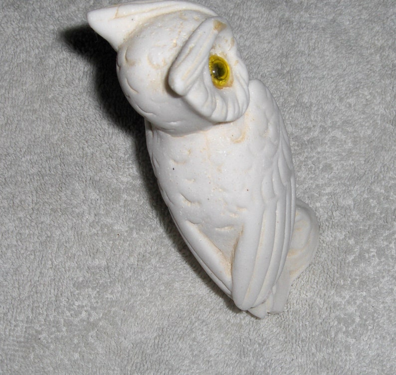 Puede incluir: Figurita de b&uacute;ho de cer&aacute;mica blanca con un ojo amarillo. El b&uacute;ho est&aacute; posado sobre una superficie blanca.