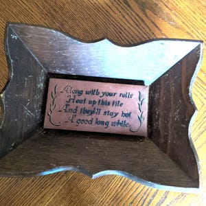 May include: A wooden framed decorative tile with the text "Along with your rolls, Heat up this tile, And they'll stay hot, A good long while." The frame is dark brown with a light blue accent.