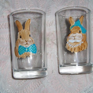 May include: Two clear glass tumblers with painted images of rabbits. One rabbit is wearing a blue bow tie and the other is wearing a blue headband with white polka dots and a pearl necklace.